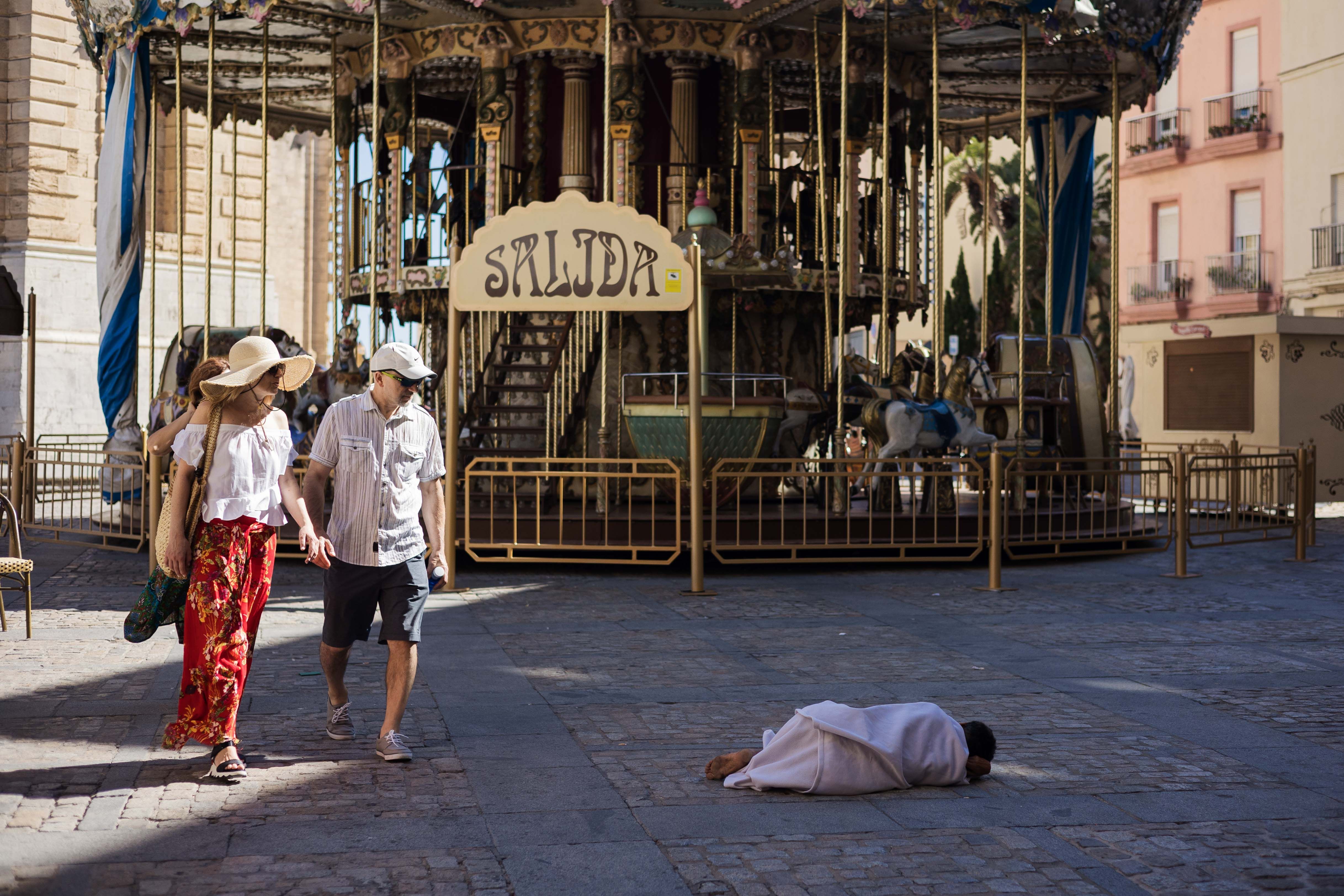 Imagen de archivo con una persona sin hogar tumbada en la plaza de la Catedral en Cádiz, ante la mirada de unos turistas. Andalucía vuelve a protagonizar el ranking de municipios más pobres.