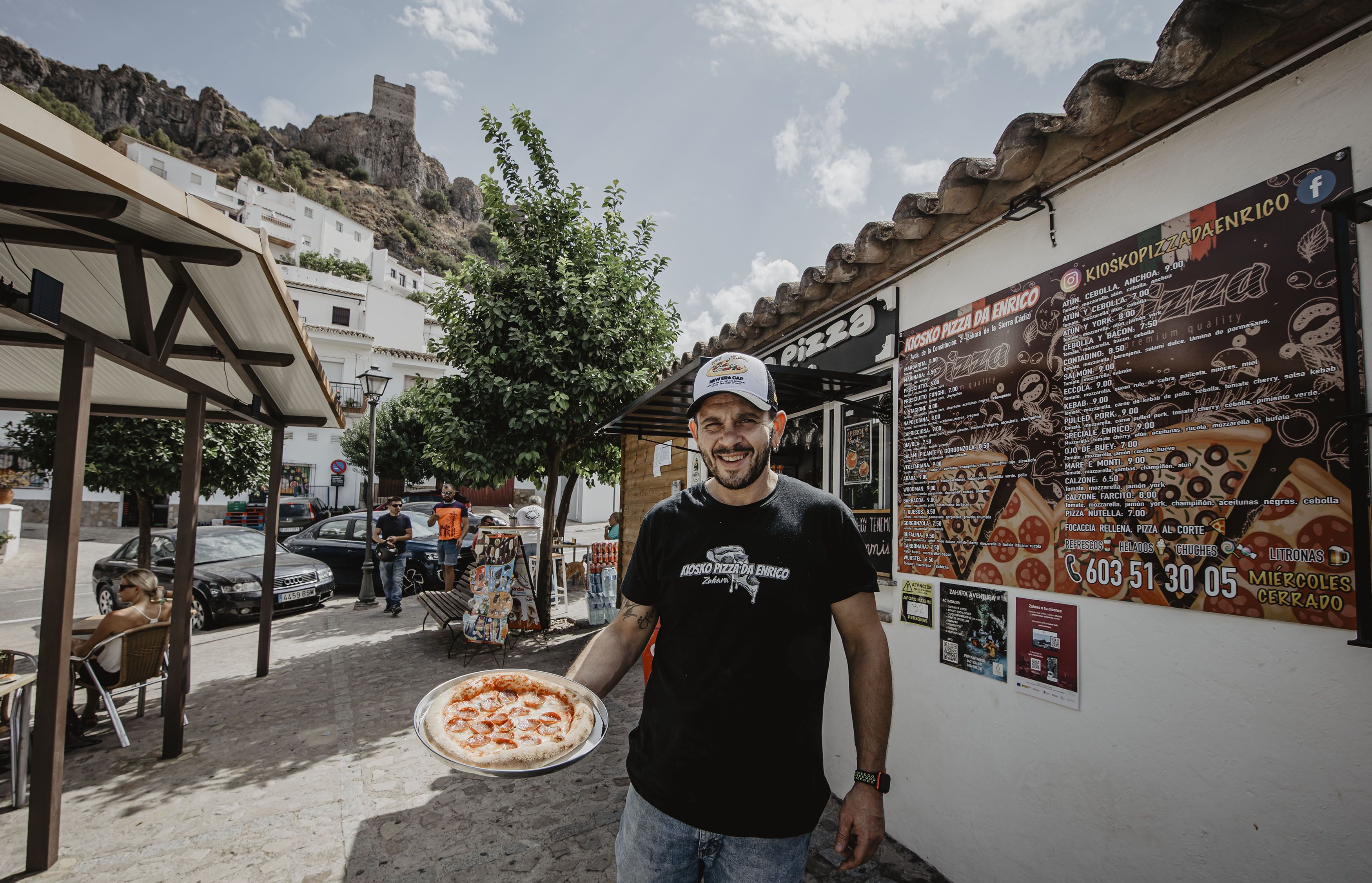 Enrico Granieri muestra una de sus pizzas artesanas, con su quiosco al fondo y el castillo de Zahara de la Sierra, donde ha echado raíces junto a su familia, en lo más alto.
