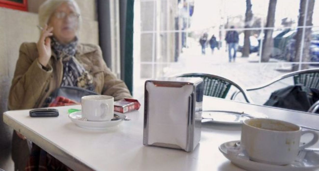 Una señora fuma mientras se toma un café en una terraza cerrada.