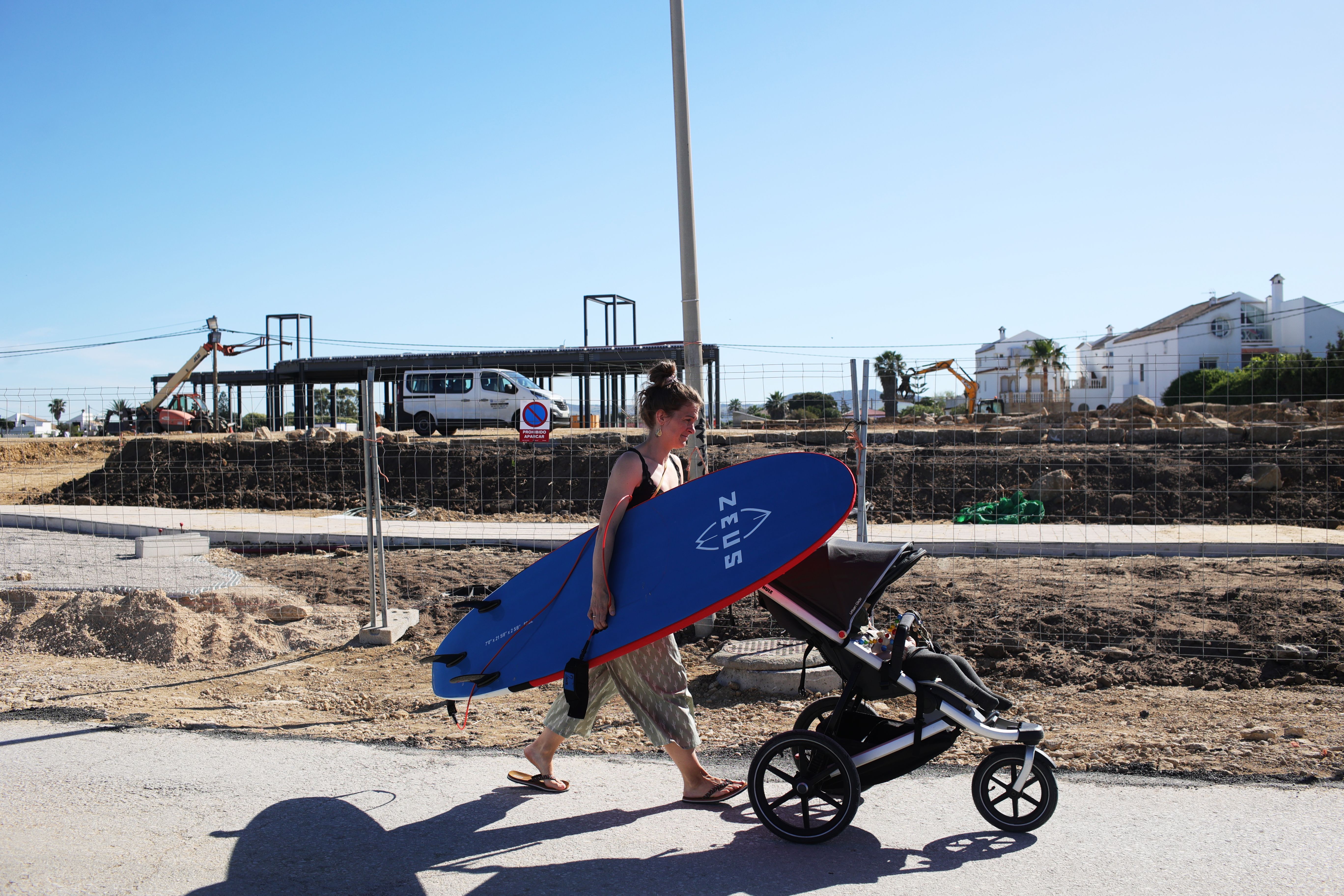 Una mujer pasa con una tabla de surf y un bebé por delante de la obra del 'beach club' Valhalla, en El Palmar.