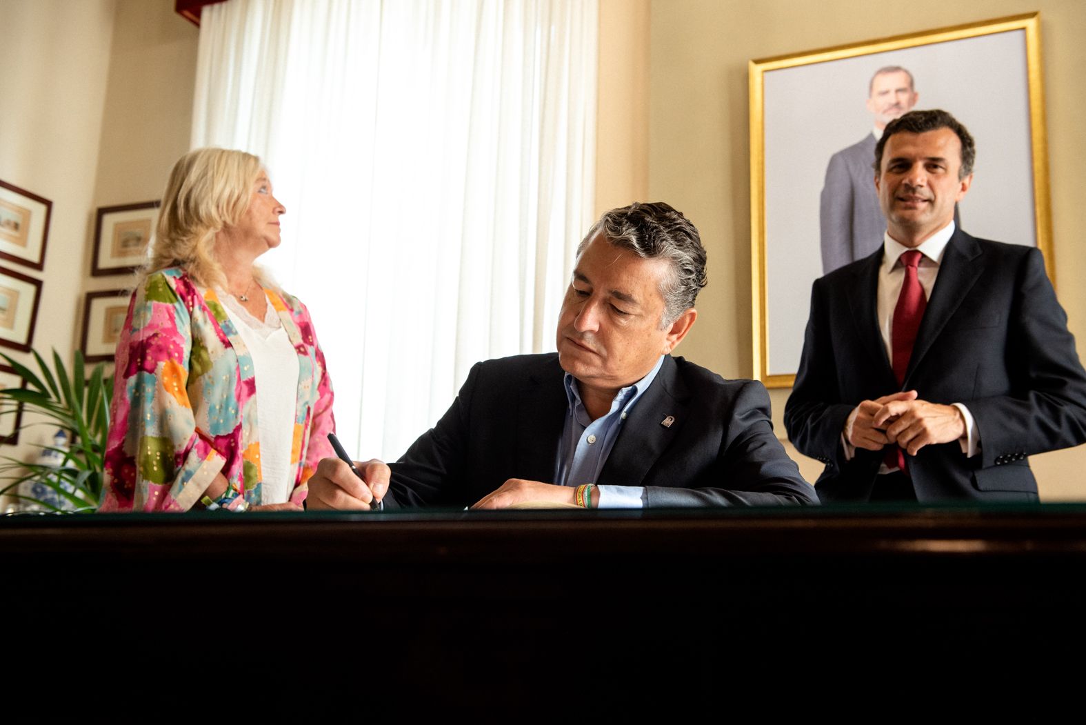 Sanz firma en el Libro de Honor mientras Bruno García le observa y Colombo mira el retrato de Felipe VI.