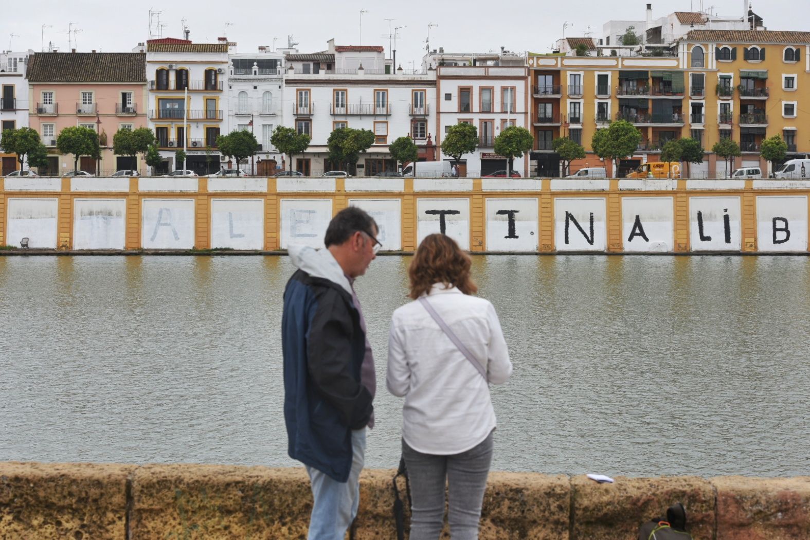 Dos personas junto al río, frente a la pintada pro Palestina en calle Betis.