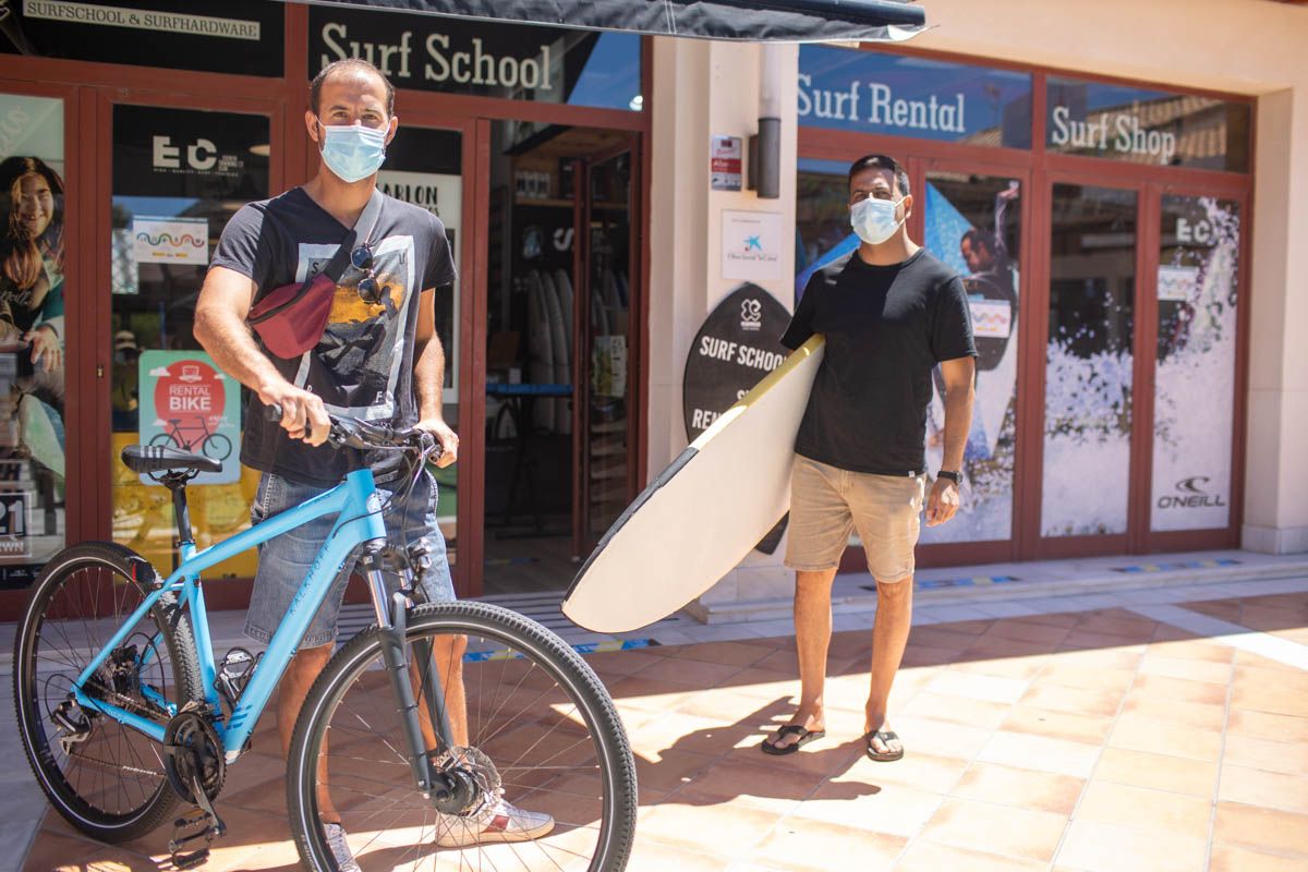 Fran, tabla de surf en mano, y Javi, con una bici, a las puertas de la tienda y escuela Esencia, en el Novo Sancti Petri, en días pasados. FOTO: JUAN CARLOS TORO