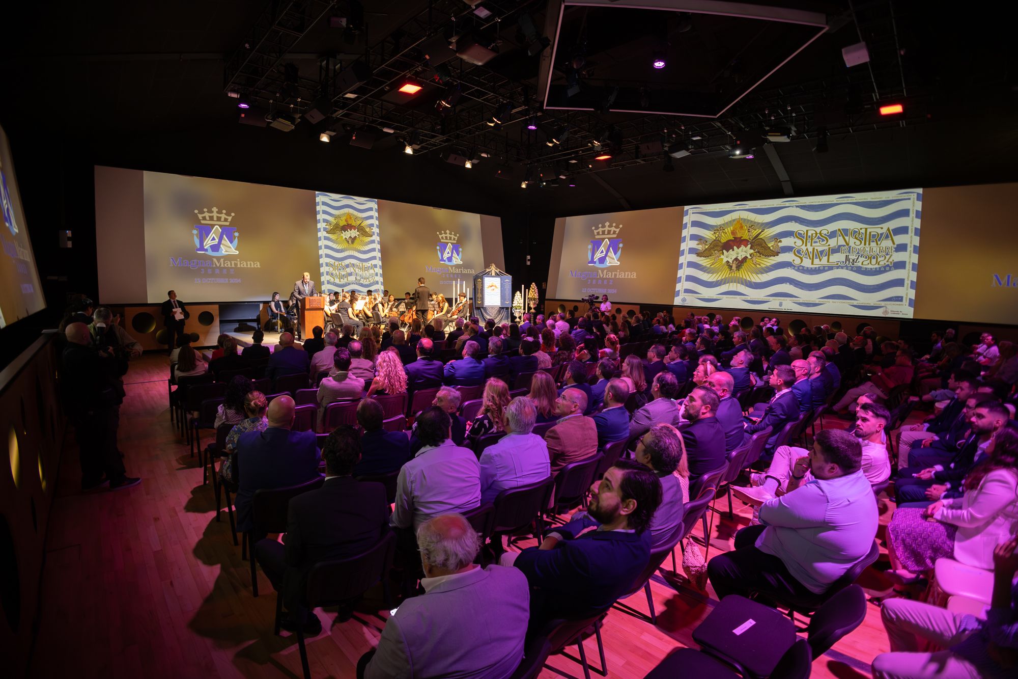 Imagen del público asistente con las pantallas del salón proyectando imágenes. 