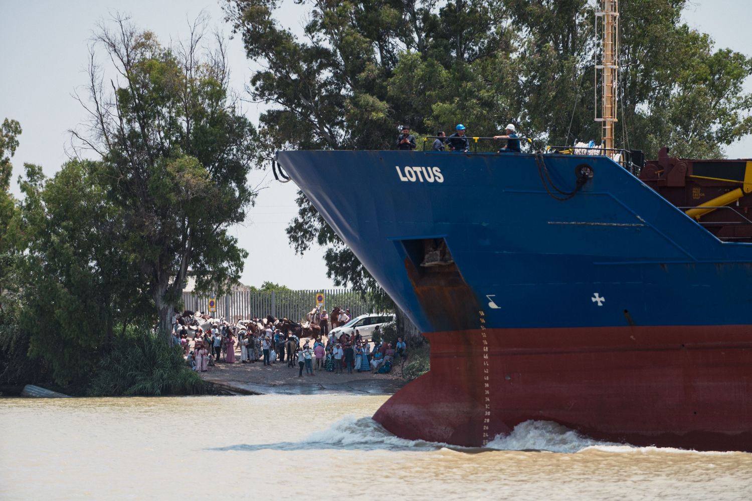 Un carguero pasa por delante de hermanos de la Hermandad del Rocío de Alcalá de Guadaíra, que esperan la barca al otro lado de Coria, para cruzar y seguir su peregrinación.
