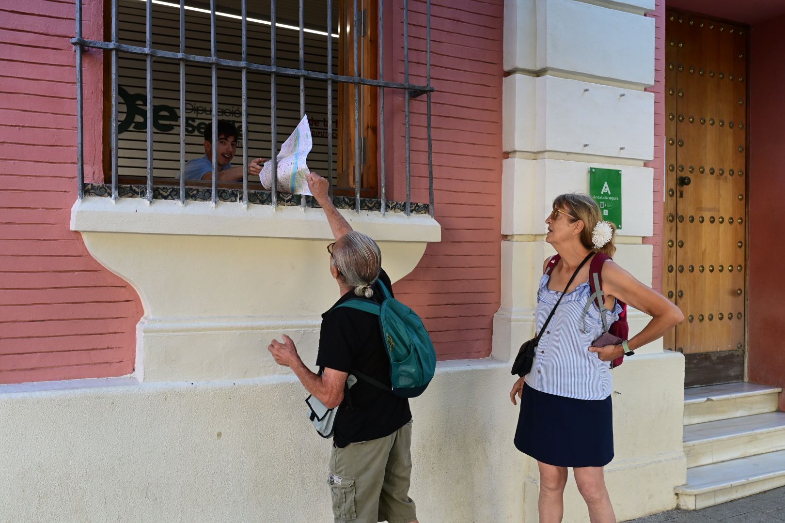 Un trabajador entrega un mapa de Sevilla por la ventana a dos turistas.