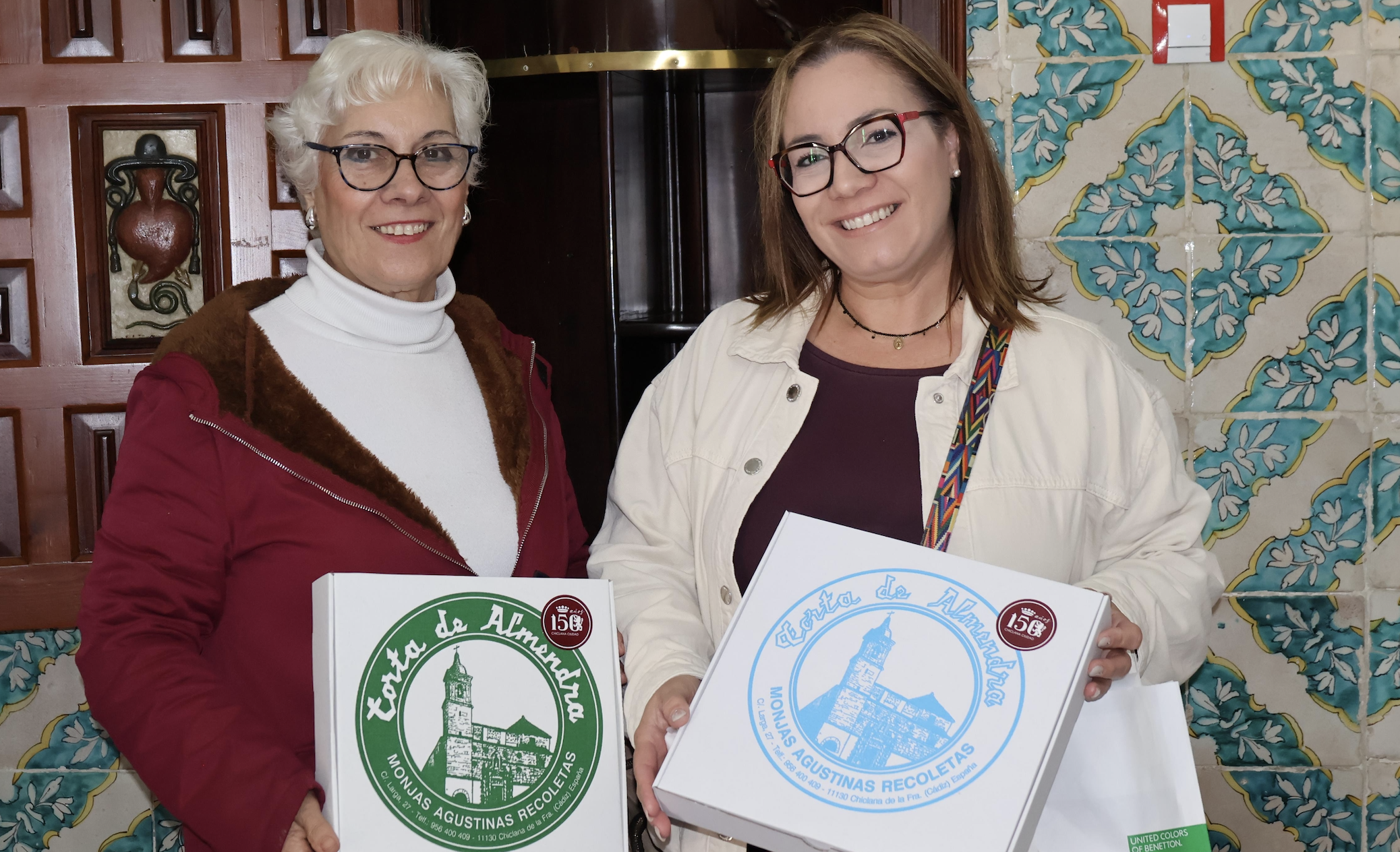 Dos señoras, ante el torno del convento, muestran las cajas de las tortas con el logo de la efeméride.