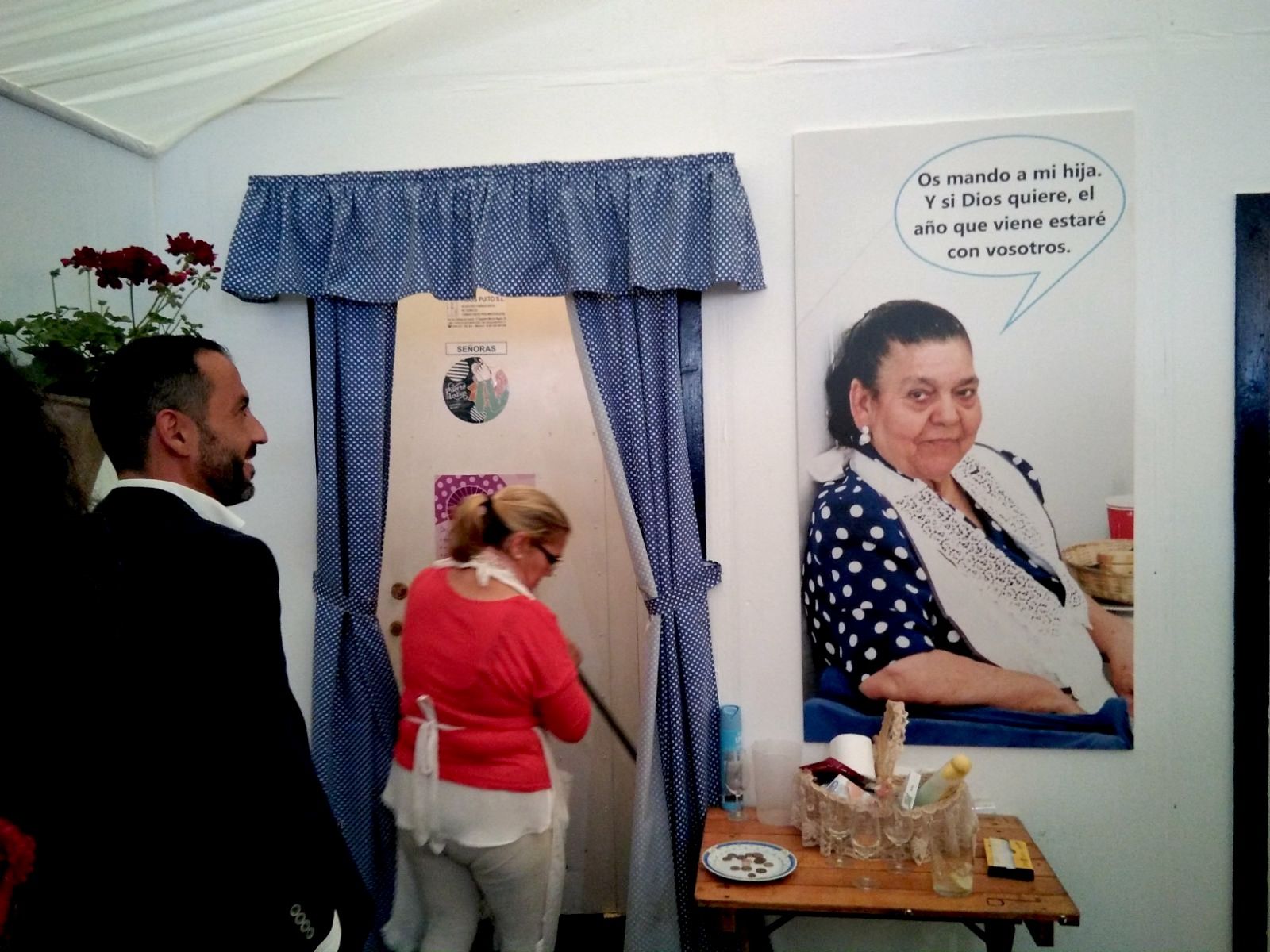 El cartel de Pilar López 'La Chana' en la caseta La Cantarera, con su hija Inmaculada limpiando el baño. FOTO: CLAUDIA GONZÁLEZ ROMERO.