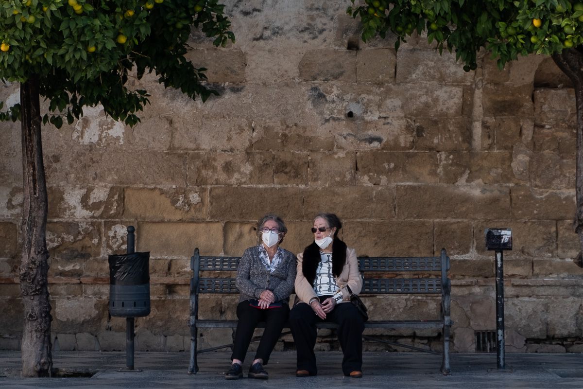Dos mujeres, con mascarilla, sentadas en un banco al aire libre.