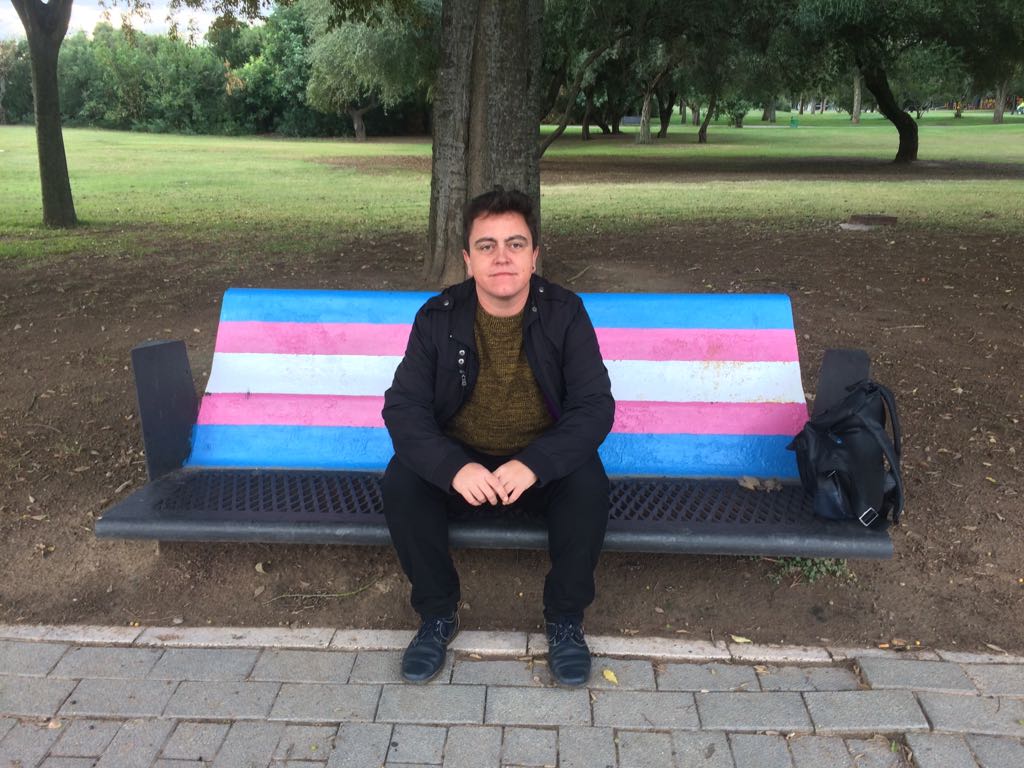 Lucas Alcázar sentado en un banco junto a la bandera trans. FOTO: LUCAS ALCÁZAR. 